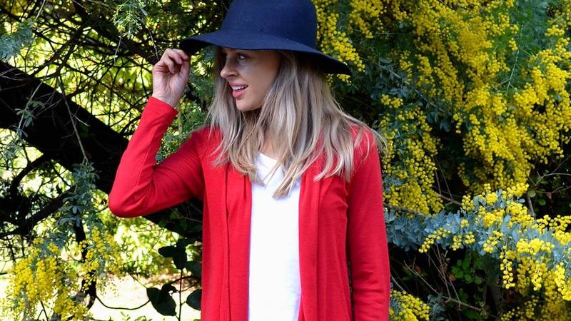 A woman in a red merino drape cardigan and navy hat in front of flowering wattle
